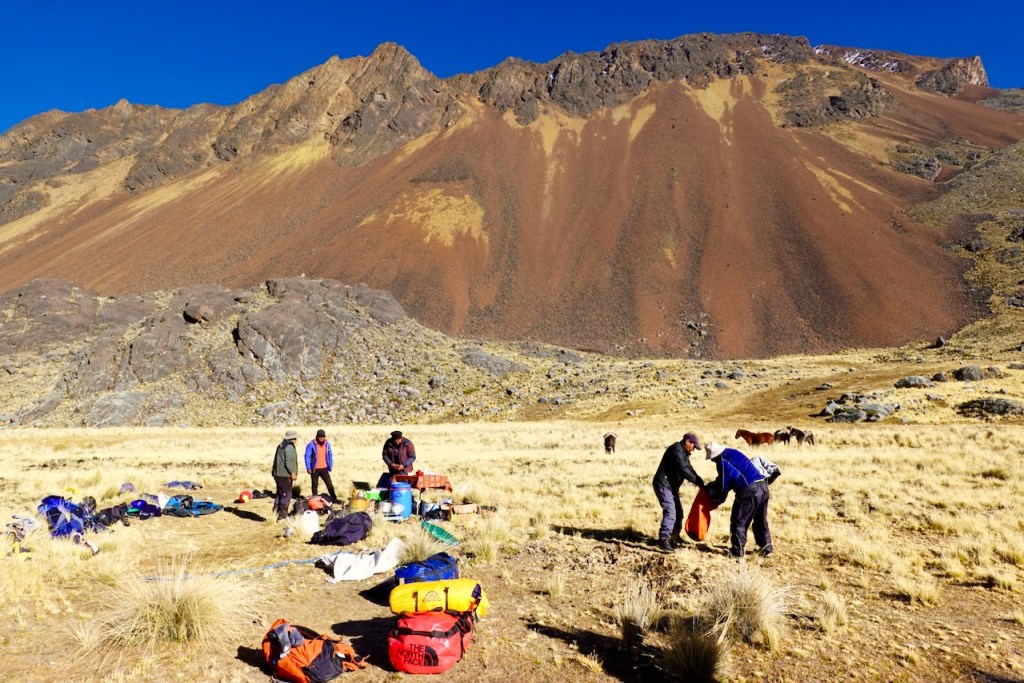 A group of people setting up camping gear in a mountainous landscape with a large rocky slope in the background.
