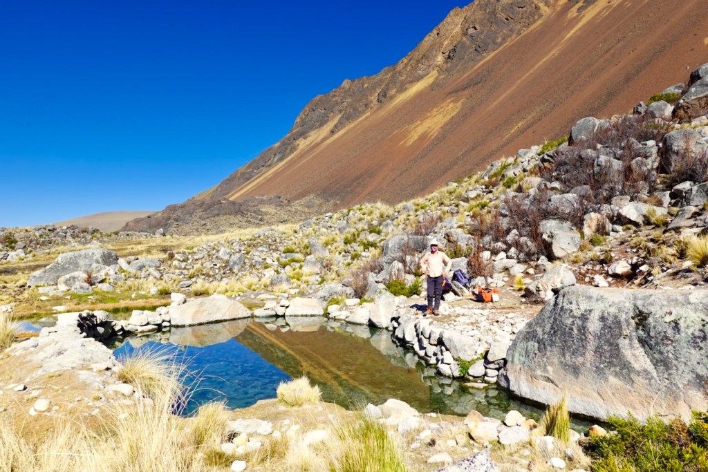 A tranquil mountain pond surrounded by rocky terrain and grass, with a clear blue sky overhead. Two people are standing near the pond, and steep mountains rise in the background.