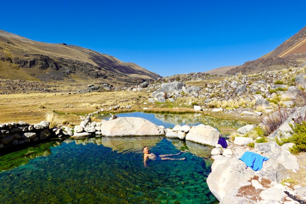A person swimming in a natural hot spring surrounded by rocky terrain and mountains under a clear blue sky.