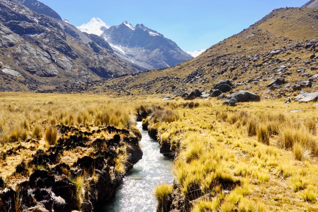 A scenic view of a river flowing through a grassy valley surrounded by towering mountains, with snow-capped peaks in the background under a clear blue sky.