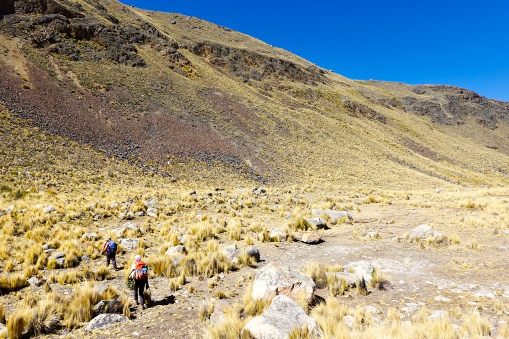 Hikers trekking through a rugged landscape with rocky terrain and sparse grass against a clear blue sky.