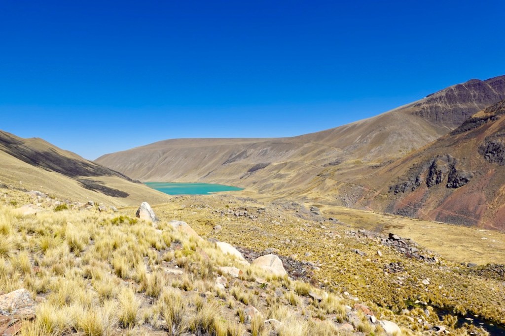 A panoramic view of a mountainous landscape featuring a turquoise lake surrounded by dry grassland and rocky terrain under a clear blue sky.