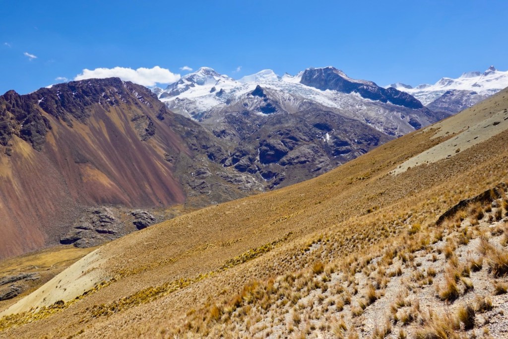 A panoramic view of mountainous terrain featuring snow-capped peaks, colorful rocky slopes, and golden grasslands under a clear blue sky.