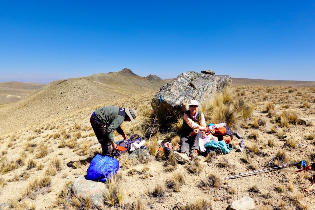 Two people preparing supplies on a rocky terrain in a mountainous area, with dry grass and clear blue sky in the background.