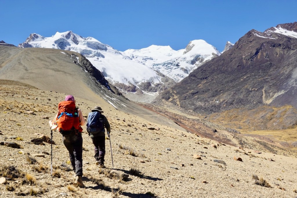 Two hikers trekking on a mountain path with snow-capped peaks in the background under a clear blue sky.