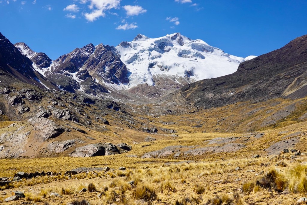 A scenic view of rugged mountains featuring snow-capped peaks under a clear blue sky, with a dry, grassy valley in the foreground.