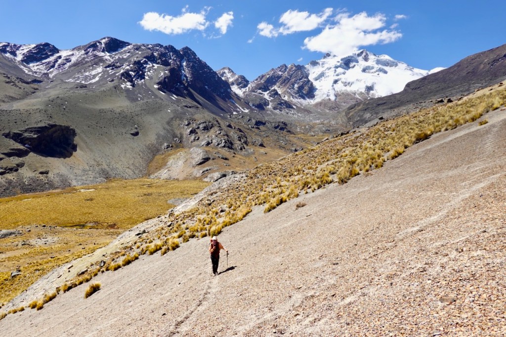A hiker traverses a rocky slope in the Andes mountains, with snow-capped peaks and clear blue skies in the background.