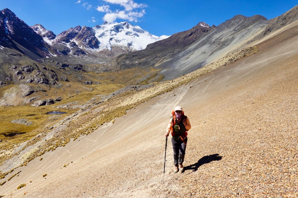 A hiker ascending a steep, rocky slope in a mountainous landscape with snow-capped peaks under a clear blue sky.