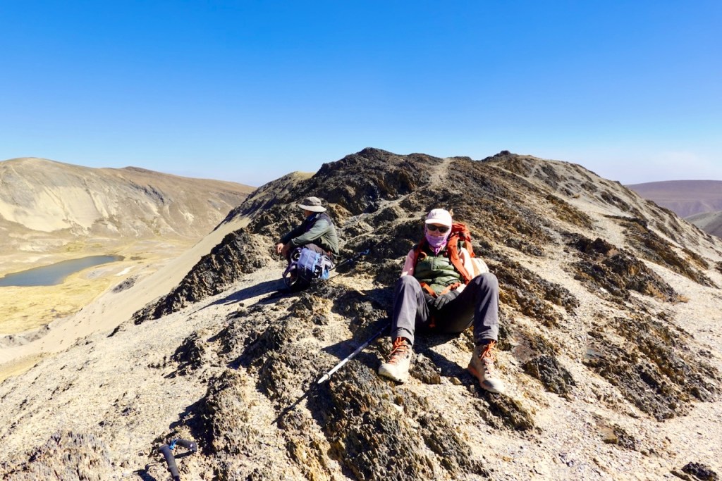 Two hikers resting on a rocky mountain peak with a clear blue sky in the background.