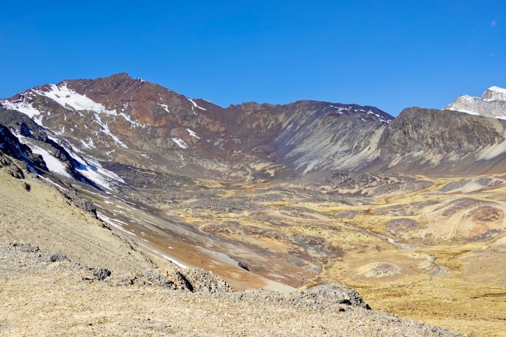 A panoramic view of a rugged mountainous landscape featuring snow-capped peaks and arid terrain under a clear blue sky.