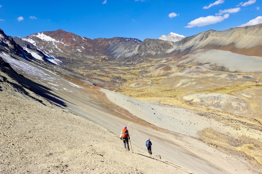 Two hikers ascend a large, rocky slope in a mountainous landscape with patches of snow and varied terrain under a blue sky.