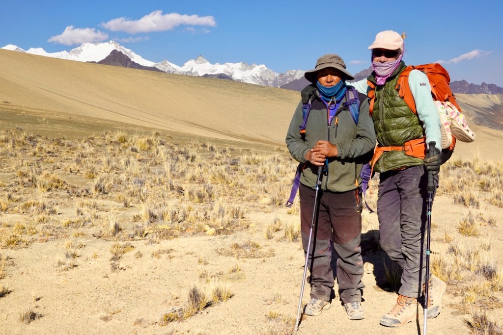Two hikers standing on a mountainous terrain with snow-capped peaks in the background, wearing colorful outdoor gear and holding trekking poles.