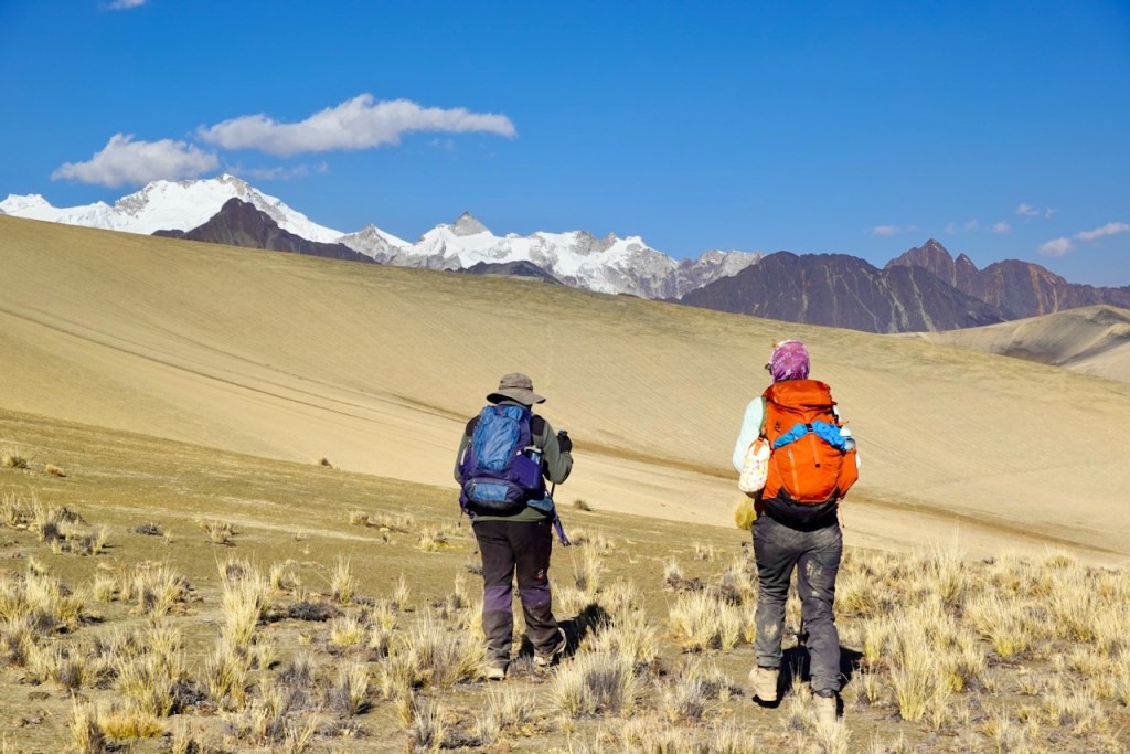 Two hikers walking on a barren landscape with grass, mountains, and snow-capped peaks in the background under a clear blue sky.