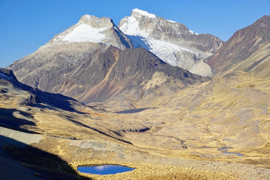 A panoramic view of a mountainous landscape featuring rugged peaks, some with snow caps, and a dry, golden valley with a small glacial lake in the foreground.