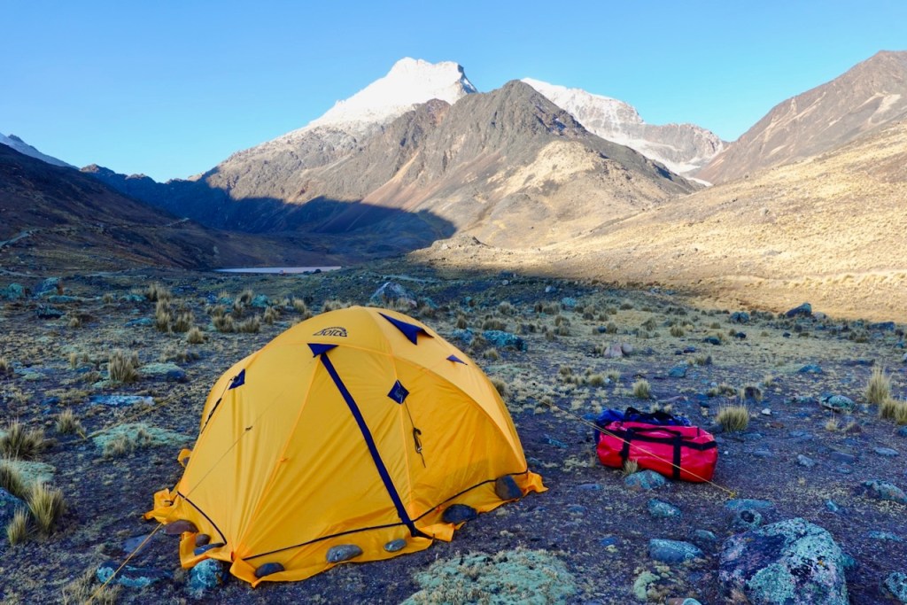 A bright yellow camping tent set against a mountainous landscape, with rocky terrain and sparse vegetation in the foreground, and snow-capped peaks in the background.
