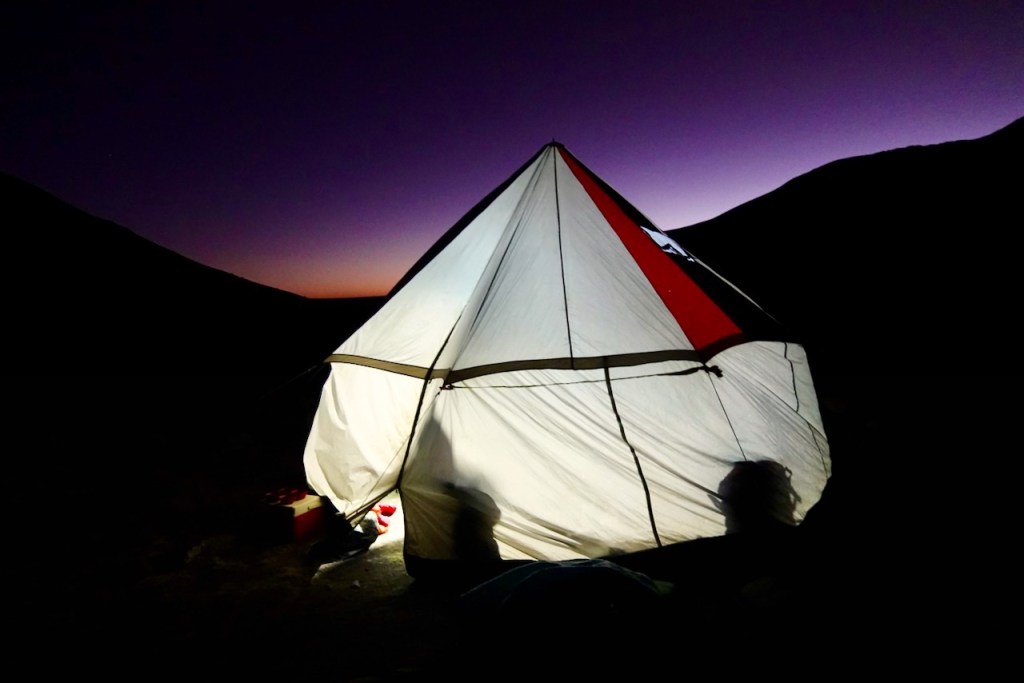 A tent illuminated at dusk, set against a backdrop of dark mountains and a gradient purple sky.