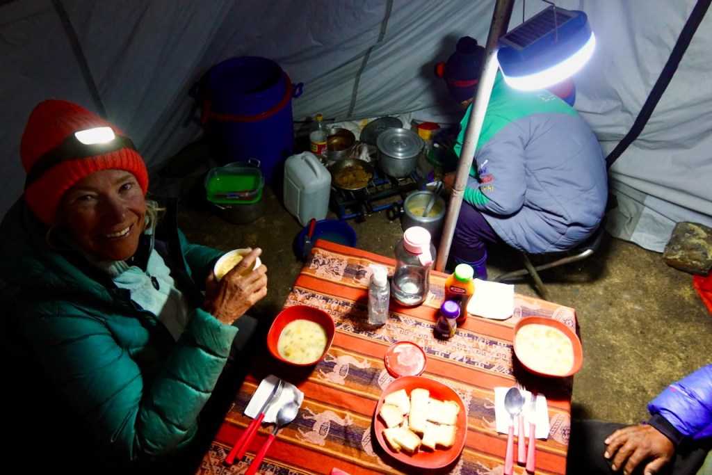 A woman with a headlamp is enjoying a bowl of soup inside a tent, with a table set for dinner and another person cooking in the background.