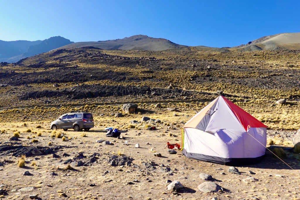 A camping tent set up in a rocky landscape with sparse vegetation, surrounded by mountains under a clear blue sky. A parked vehicle and camping gear are visible nearby.