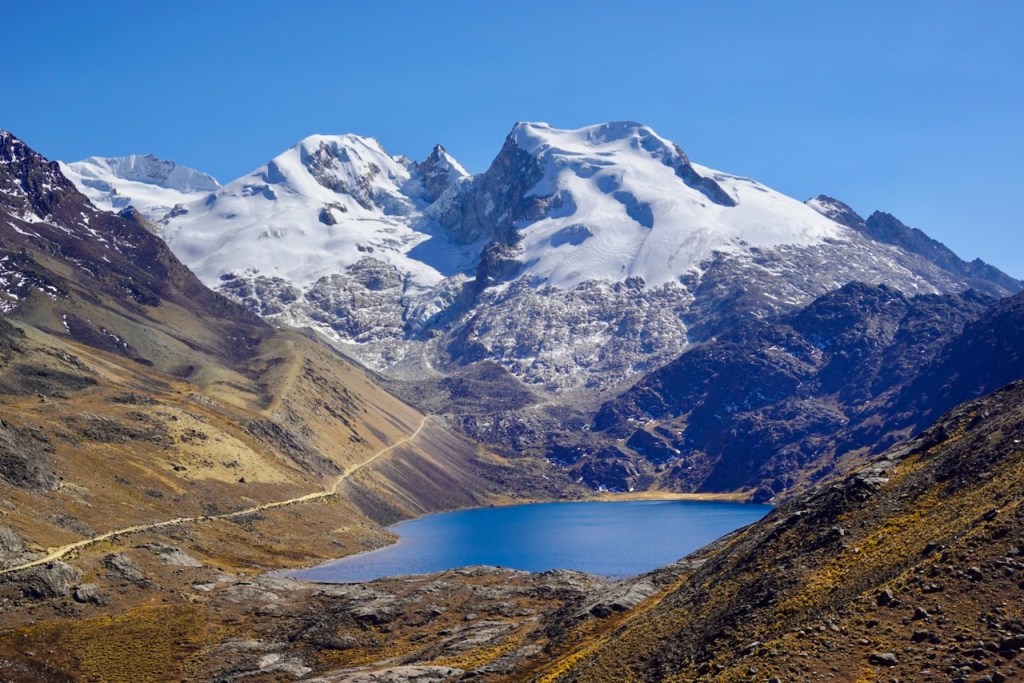 A panoramic view of a clear blue lake surrounded by rugged mountains and snow-capped peaks under a bright blue sky.