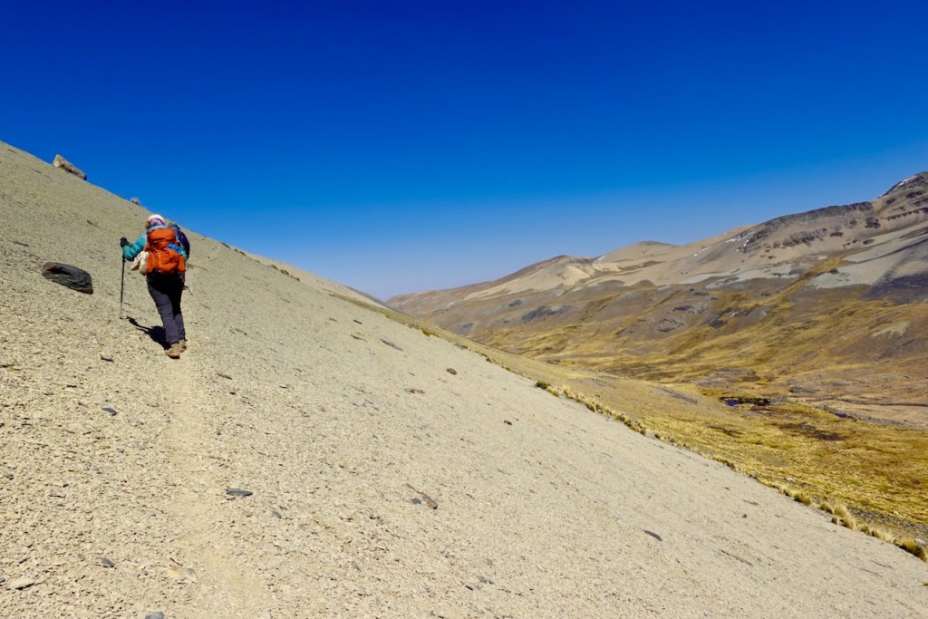 A hiker ascends a steep, sandy slope in a mountain landscape under a clear blue sky.