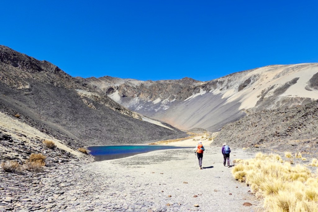 Two hikers walking along a rocky path beside a vibrant blue lake, surrounded by mountains and clear blue sky.