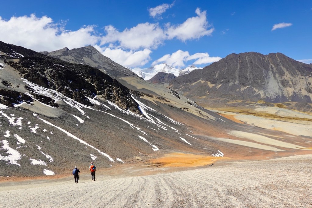 Two hikers trekking along a gravel path in a mountainous landscape with rocky terrain and snow-capped peaks under a blue sky with clouds.