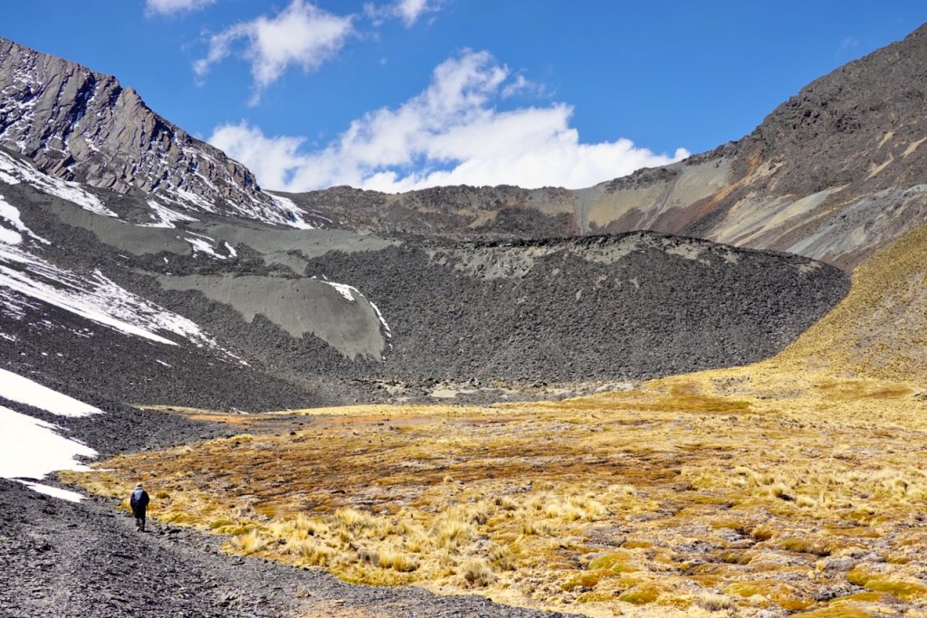 A hiker walks through a rocky landscape in a mountain valley, surrounded by steep slopes and a blue sky with scattered clouds.