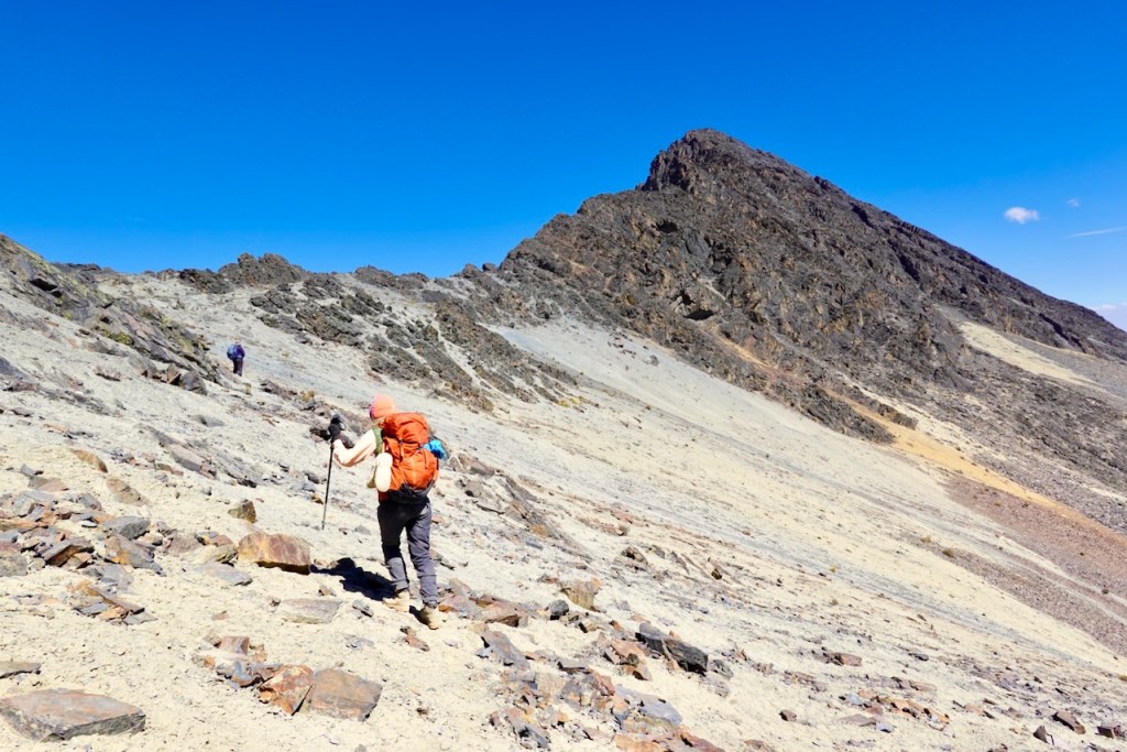 Two hikers ascend a rocky, barren slope towards a steep mountain peak under a clear blue sky.