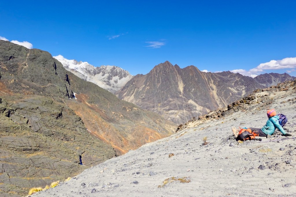 Two hikers resting on a rocky mountain slope with a panoramic view of rugged peaks and a clear blue sky.