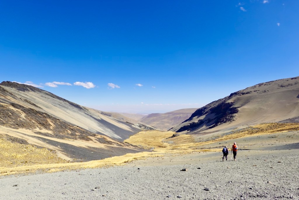 Two hikers walking through a vast, rocky valley surrounded by steep hills under a clear blue sky.