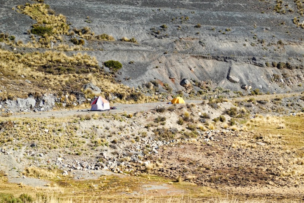 Two camping tents set up on a rocky hillside, surrounded by sparse vegetation and steep terrain.