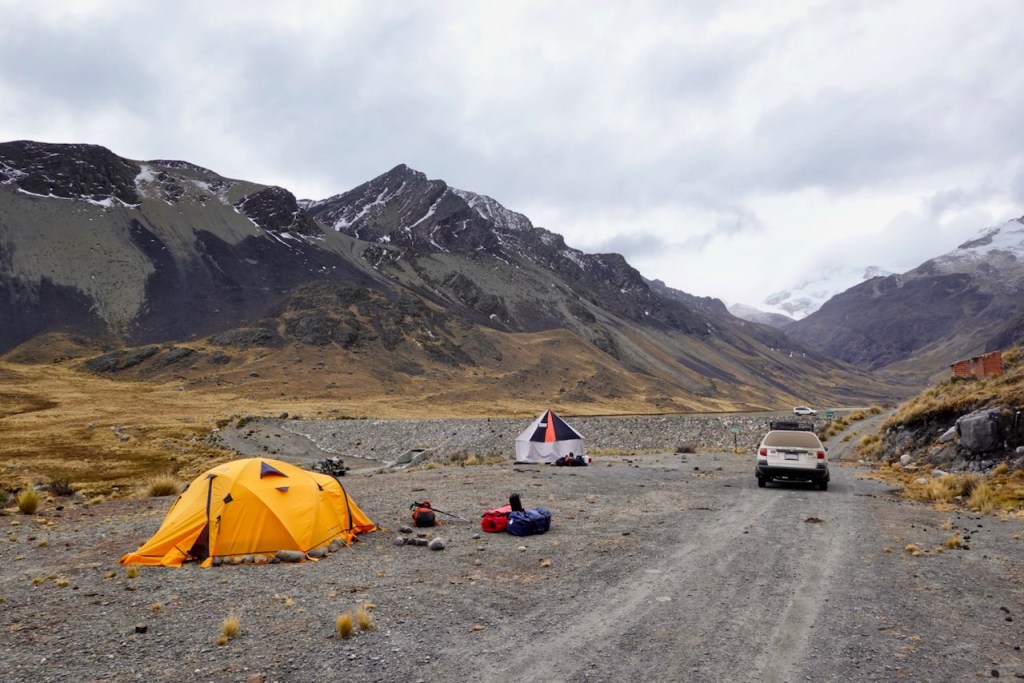 A scenic camping area featuring two tents, one orange and one white, set against rugged mountains and a cloudy sky.