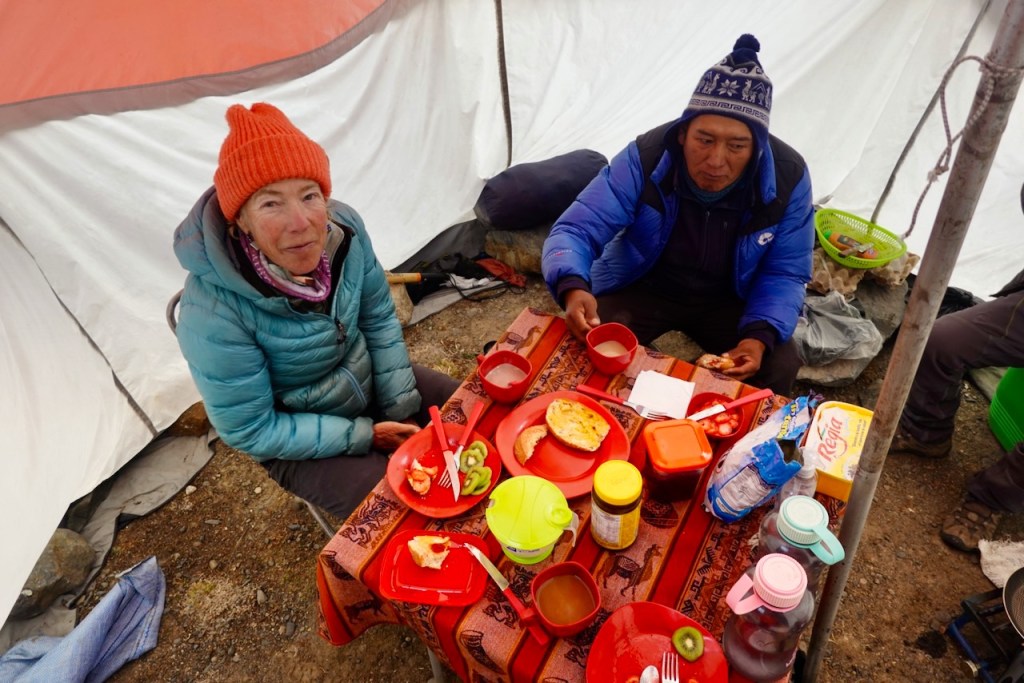 Two people sitting at a table inside a camping tent, enjoying a meal together. The table is covered with an orange patterned cloth and contains multiple plates of food, drinks, and snacks.