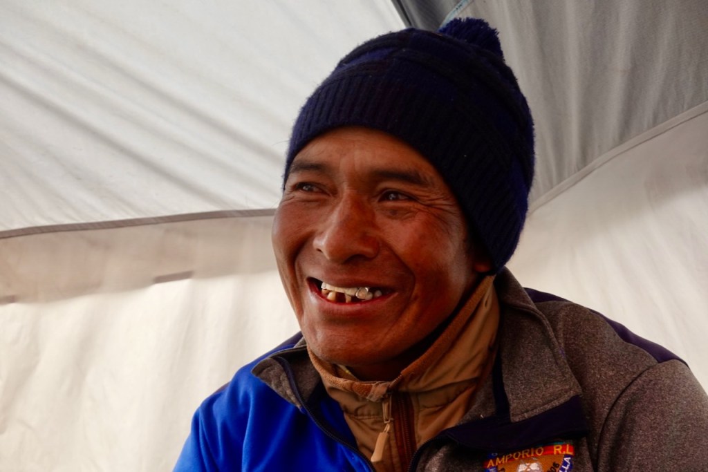 A smiling man wearing a blue hat and a gray jacket, seated inside a tent, showcasing his happy expression.