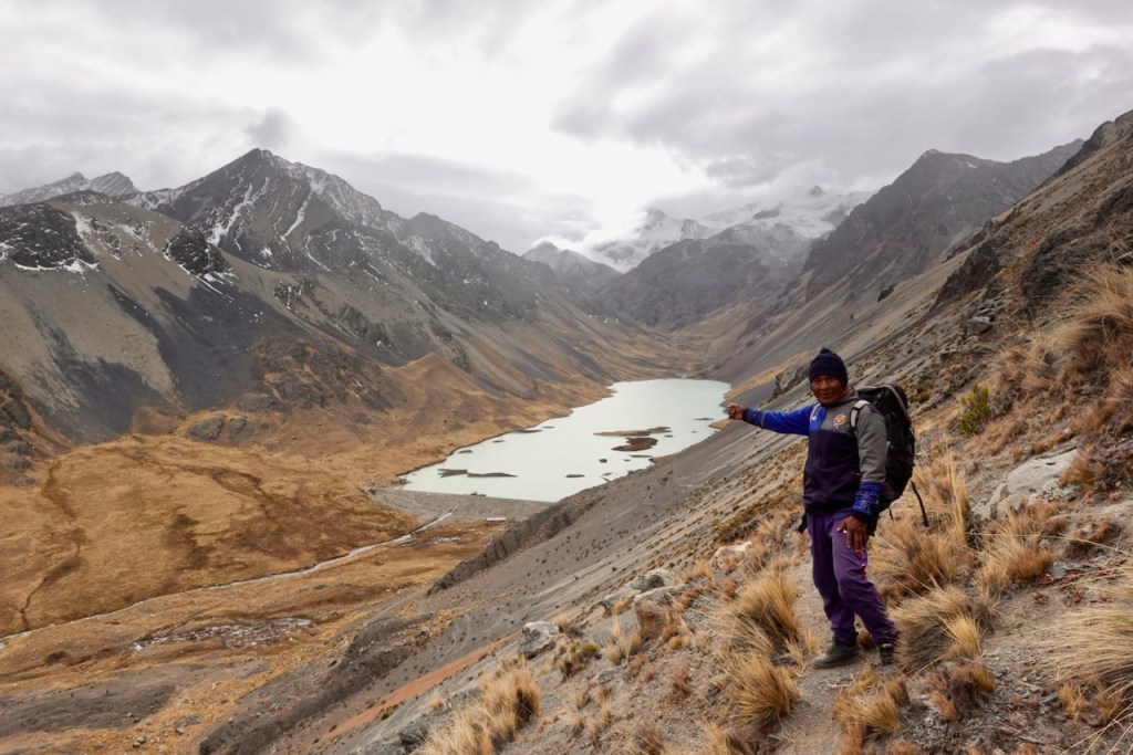 A person in a blue jacket and hat stands on a rocky slope, pointing towards a glacial lake surrounded by rugged mountains and a cloudy sky.