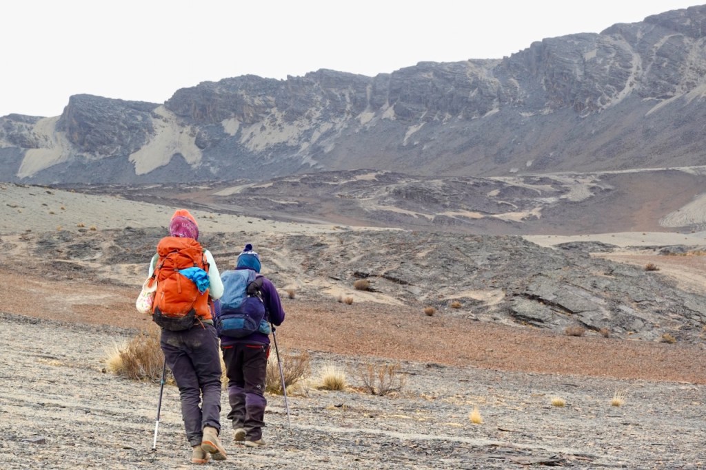 Two hikers trekking through a rocky landscape with mountains in the background.