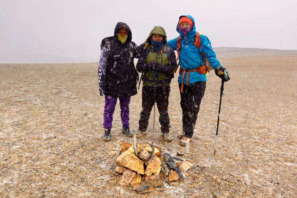 Three hikers standing together in a snowy landscape, with a small pile of stones and wood in front of them, wearing colorful winter jackets.