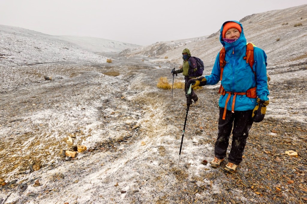 Two hikers in winter gear walking on a snowy mountain trail with rocky terrain and low visibility.