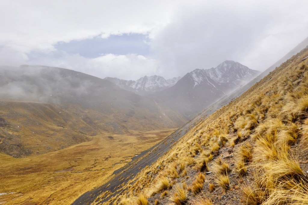 A panoramic view of a mountainous landscape with rugged peaks and a misty sky. The foreground features dry grass and a steep slope leading to a valley below, while distant snow-capped mountains are partially obscured by clouds.