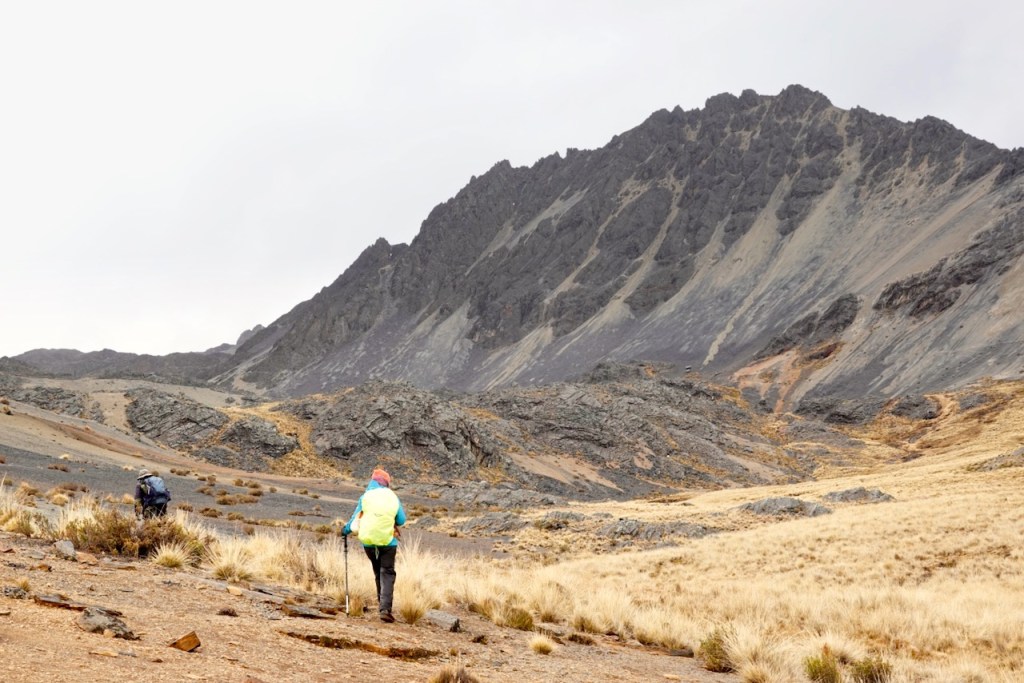 Two hikers trekking through a rocky landscape, with a prominent mountain in the background under a cloudy sky.