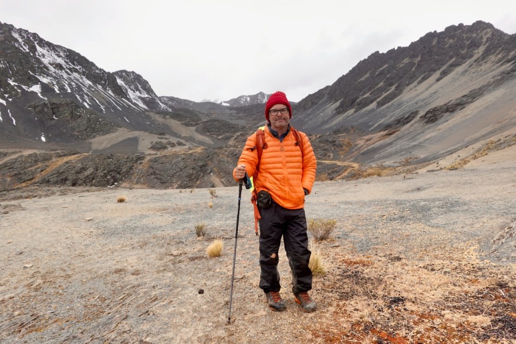 A hiker wearing an orange puffy jacket and red beanie stands on a mountainous terrain with rocky slopes and patches of snow in the background.