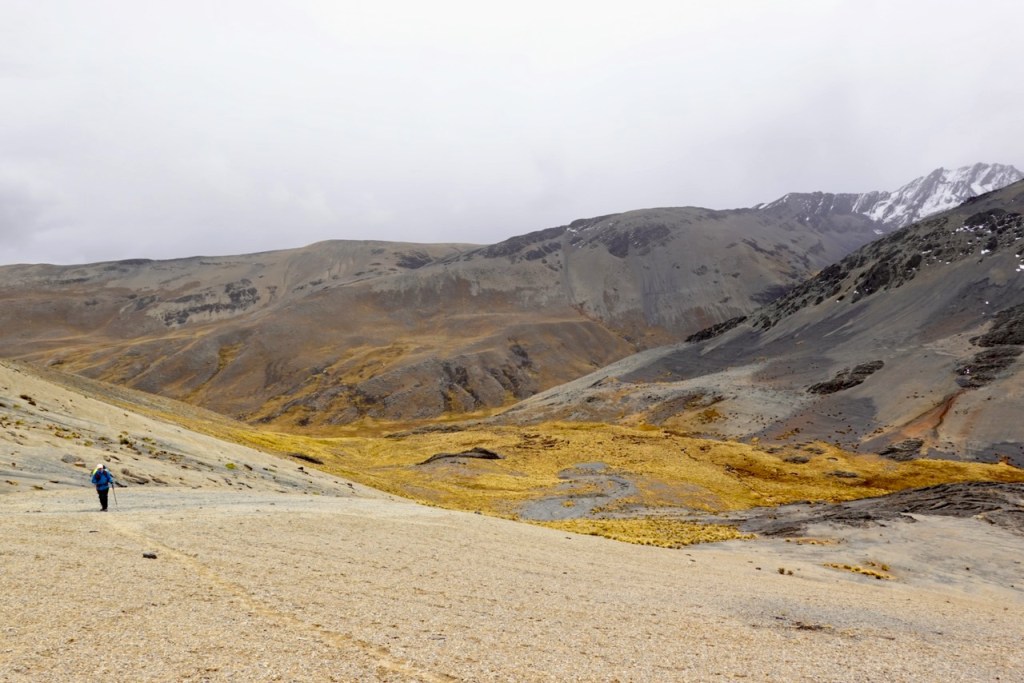 A hiker walking on a rocky terrain with mountain ranges in the background, featuring a mix of gray and yellow landscapes under a cloudy sky.