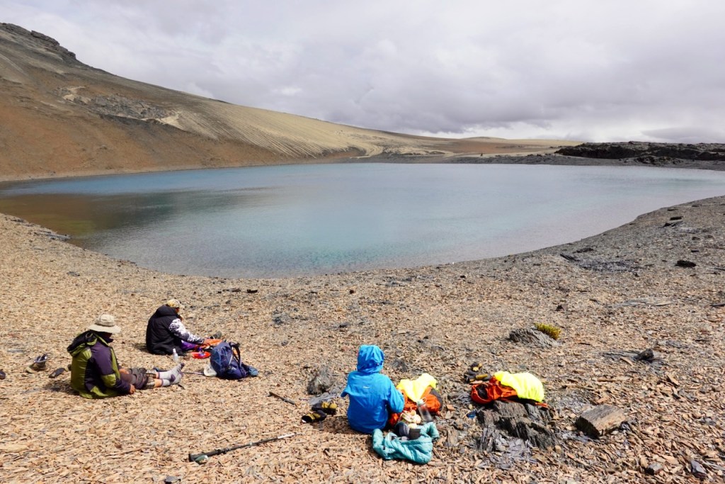 Three people sitting by a calm lake surrounded by rocky terrain and hills under a cloudy sky.