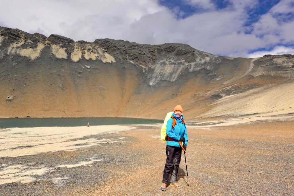 A hiker stands on a rocky shoreline near a turquoise lake, with a large mountainous slope in the background under a cloudy sky.