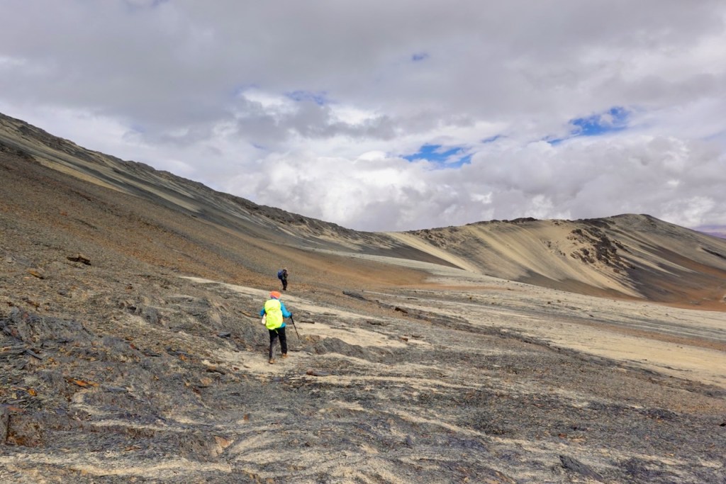 Two hikers navigating a rocky, sloping terrain under a partly cloudy sky.