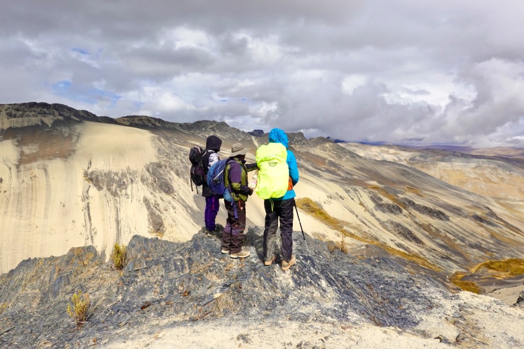 Three hikers standing on a rocky outcrop overlooking a mountainous landscape with sandy slopes and cloudy skies.