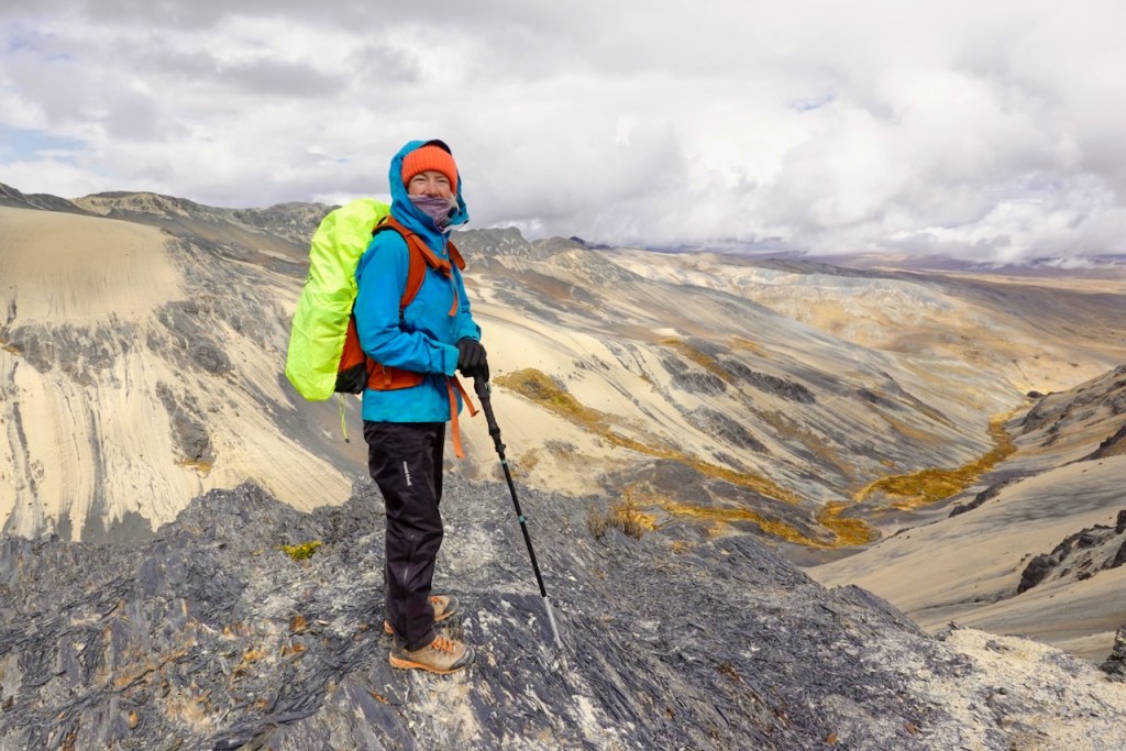 A hiker stands on a rocky outcrop, overlooking a vast landscape of mountains and valleys. The hiker is wearing a bright blue jacket, a green backpack, and has trekking poles, with cloud-covered skies in the background.