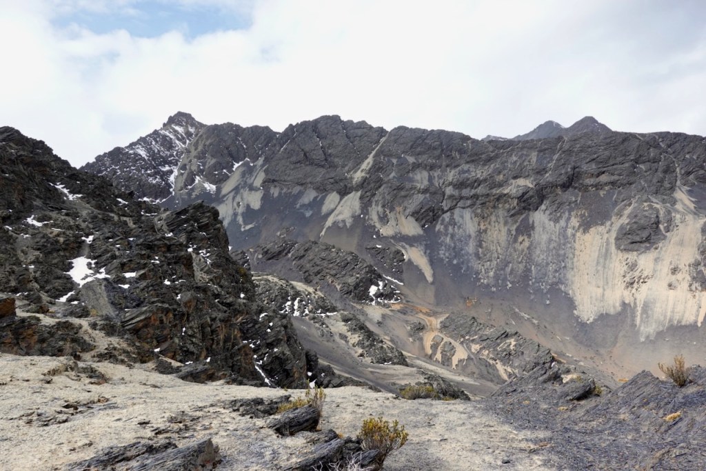 A panoramic view of rugged mountain terrain with rocky cliffs and a snow-capped peak under a cloudy sky.