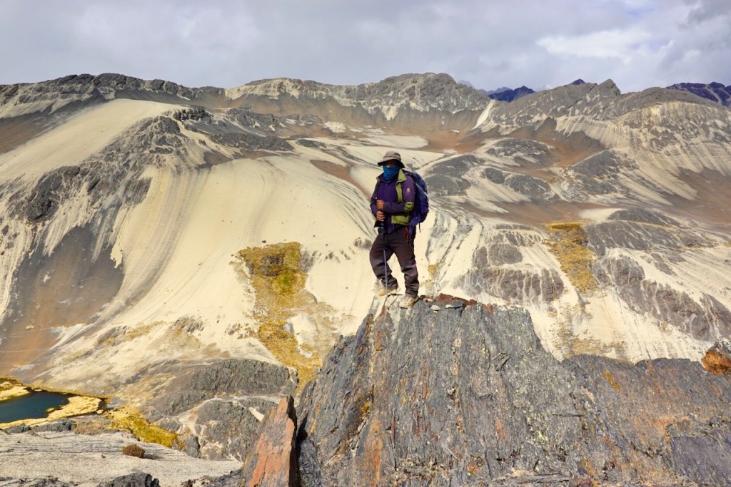 A hiker stands on a rocky summit with a vast mountainous landscape in the background, featuring layers of rock and sand.