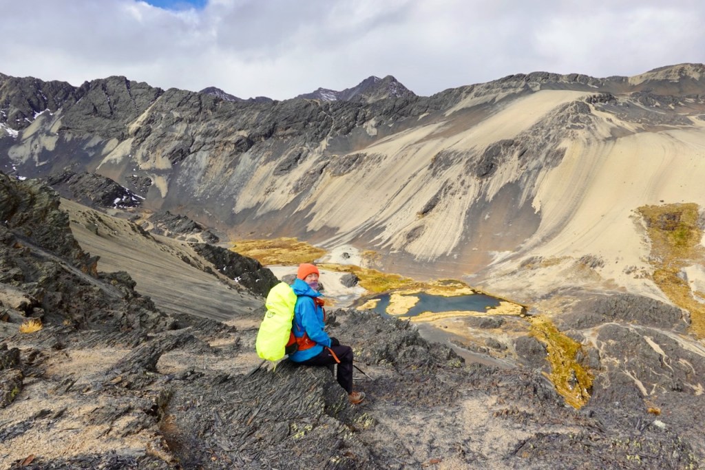 A hiker in colorful outdoor gear sits on a rocky outcrop, overlooking a scenic valley with mountains in the background and a small body of water below.